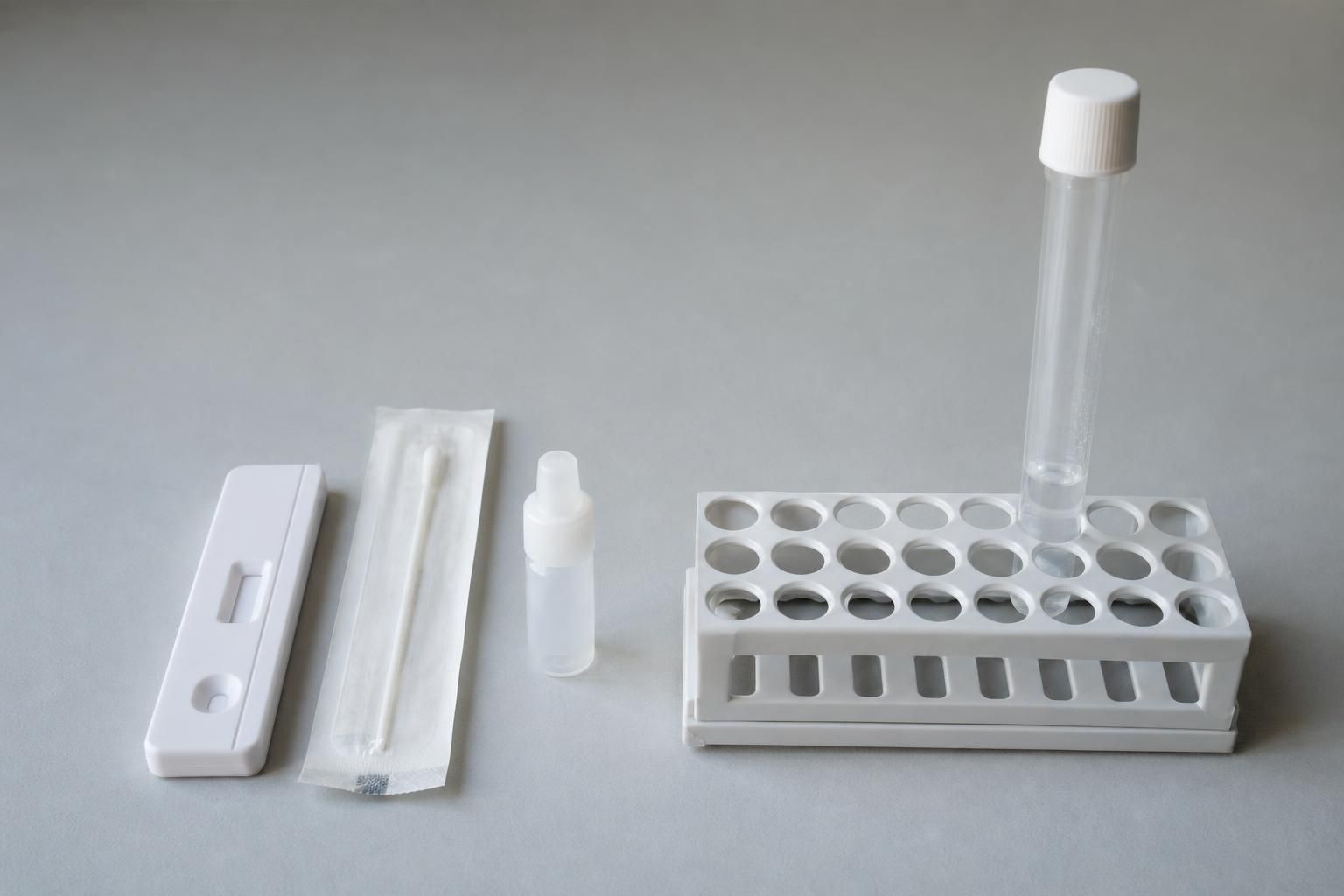 Side-by-side photo on a plain grey surface: an unbranded rapid test cassette, a sealed sterile swab, and a buffer vial on the left, and a capped, unlabeled lab specimen tube in a white rack on the right, showing the two sample-testing routes.