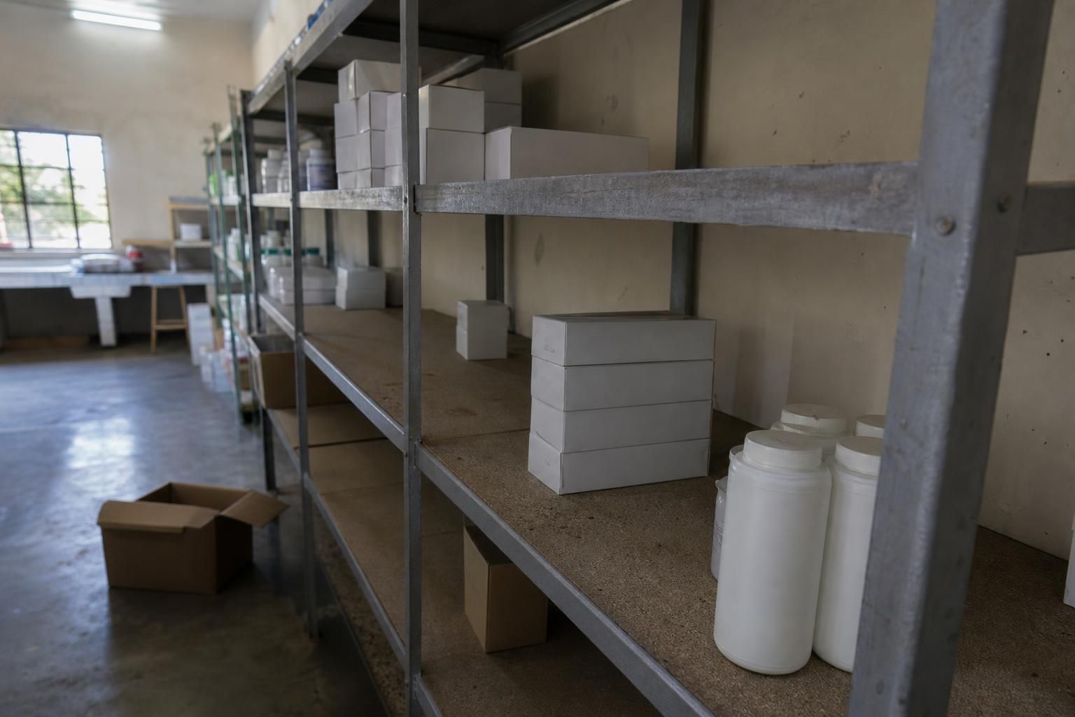 Clinic storeroom with partly empty metal shelves, showing sparse plain cartons and a few unlabeled bottles with clear gaps where supplies have run out.