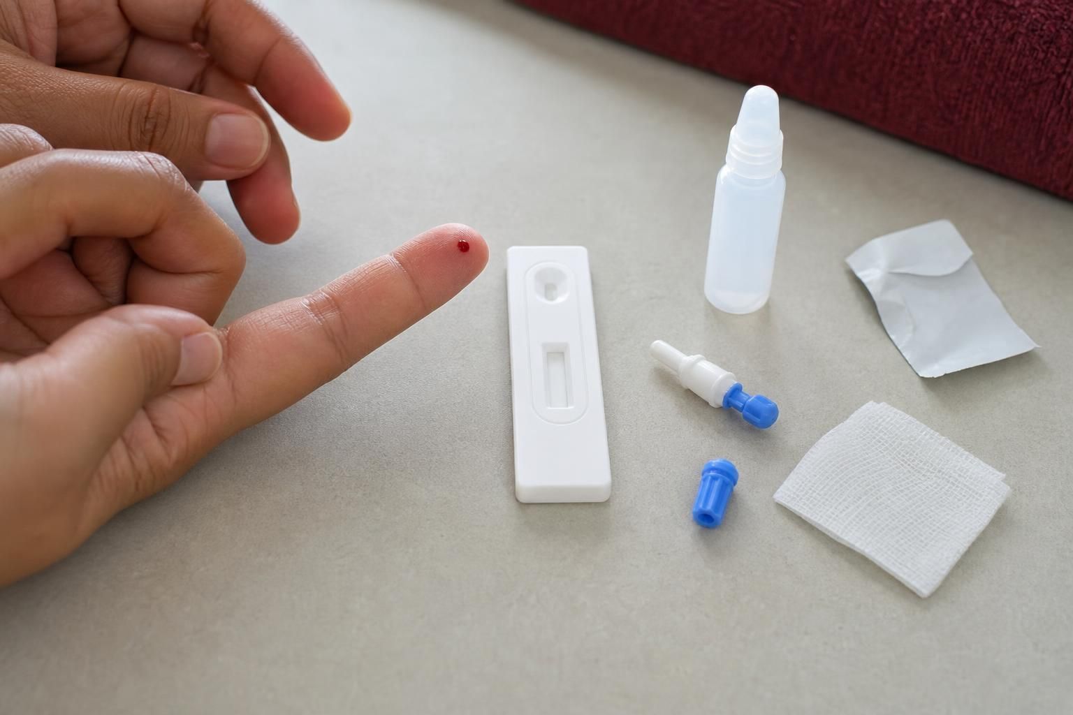 An adult's hands with one small drop of blood on a fingertip, beside an unbranded rapid test cassette with a blank result window, a lancet, an alcohol prep pad, and a buffer dropper bottle on a countertop.