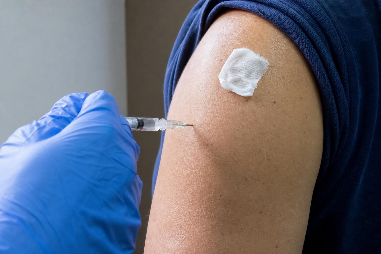 Close-up of a gloved hand giving an intramuscular hepatitis B vaccine injection into the upper outer arm of an adult, with an alcohol swab on the skin beside the injection site.