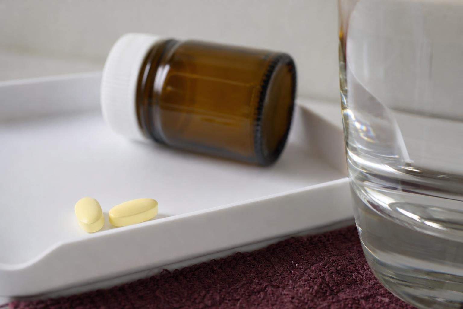 Two plain, unmarked pale yellow tablets on a white pharmacy tray, with an unlabeled amber vial and a glass of water beside them.