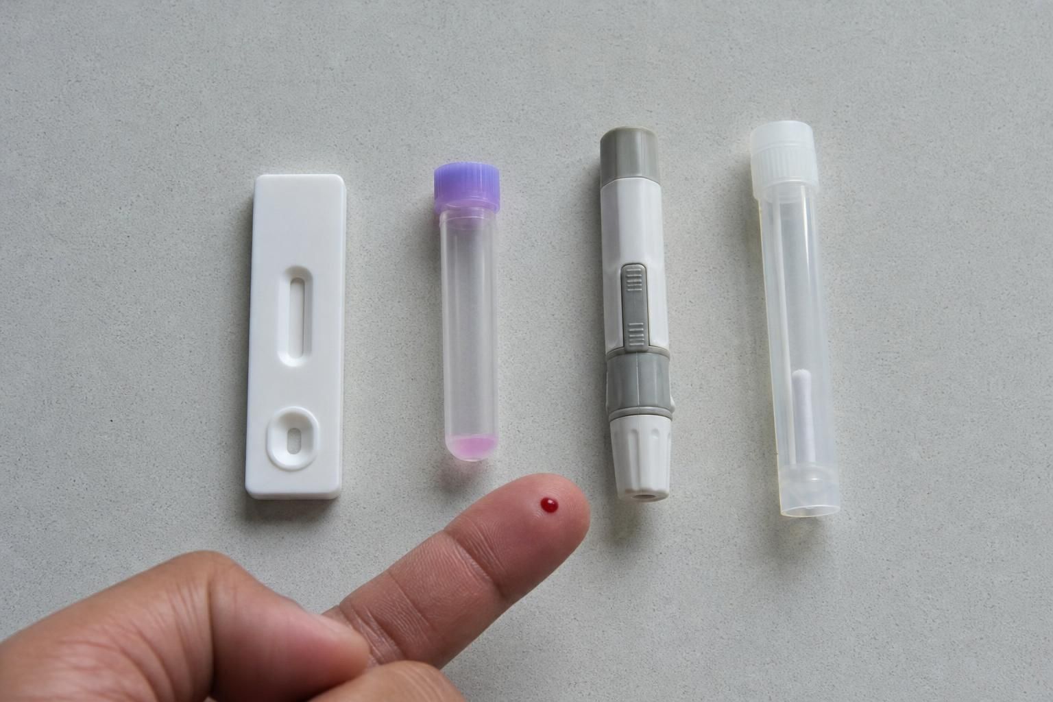 Overhead view of four unbranded testing supplies on a plain grey counter: a rapid test cassette, a blood collection tube, a lancet and a sealed oral swab tube, with one fingertip showing a small bead of blood at the lower edge.