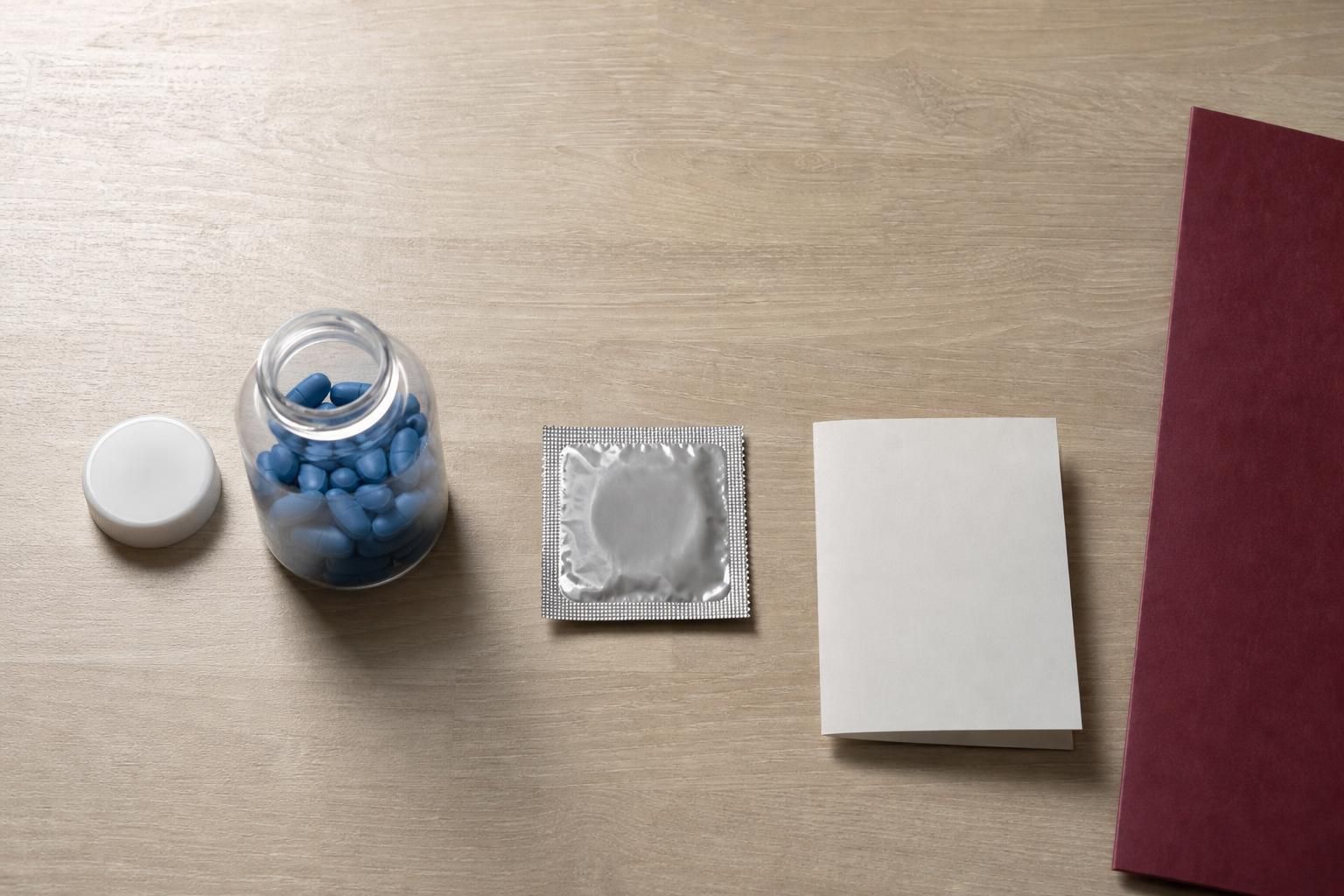 Overhead view of a pale wooden table with an unlabeled bottle of blue tablets, a sealed condom packet, and a blank folded card, showing everyday objects connected to HIV prevention decisions.